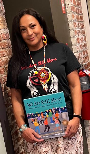 Amelia Martinez stands holding a copy of the picture book, We Are Still Here!