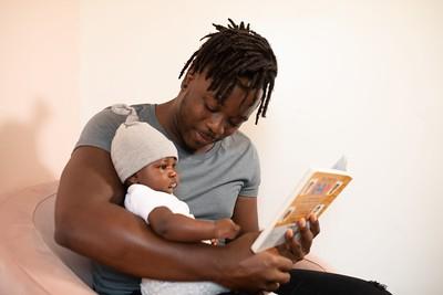 African American with dreads reading to an infant on their lap. The infant is wearing a cap and reaching for the book.