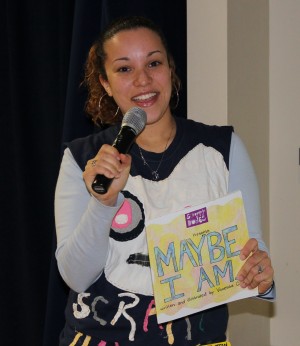 Vanessa Campos, an author is featured holding a microphone in her right hand and a copy of her book, Maybe I Am,in her left hand.