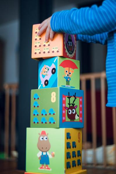 child hands and arms are visible as they place a block on a tower of blocks. The child is wearing a long sleeved blue stripped shirt. Each side of the blocks feature an animal character or number with objects that can be counted for that number.