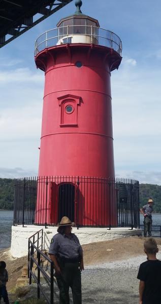 The Little Red Lighthouse under the George Washington Bridge. The lighthouse is surrounded by a black fence. Two park rangers are present and one child is looking at the lighthouse.